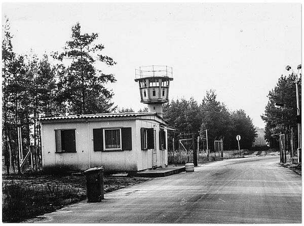 "Dienstkomplex Freienbrink" der Stasi: Zufahrtstor (o. D.); Quelle: BArch, MfS, GH, Nr. 16/84, Bd. 1, Bl. 54, Bild 2 Blick die Zufahrtsstraße zum Stasi-Gelände in Freienbrink hinunter: links der Straße ein hüttenartiges Wärterhäuschen, dahinter ein Wachturm.