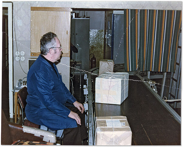 "Dienstkomplex Freienbrink" der Stasi: Mitarbeiter der Abt. M (Postkontrolle) bei der Überprüfung von westdeutschen Paketen (nach 1984); Quelle: BArch, MfS, Abt. M, Fo, Nr. 30, Bild 1 Innenraum: ein von seiner rechten Seite fotografierter etwa fünzig Jahre alter Stasi-Mitarbeiter sitzt mit einem blauen Arbeitskittel bekleidet vor einem kleinen Förderband auf dem Pakete vor ihm entlangbefördert werden.