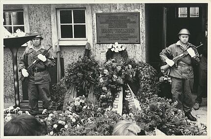 Zwei uniformierte und bewaffnete Soldaten stehen in einem Hof neben einer Plakette, die an die dort stattgefundene Ermordung Ernst Thälmanns erinnert. Vor und neben ihnen ist eine Vielzahl von Blumengestecken und Blumenkränzen.