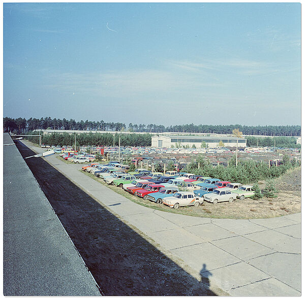 "Dienstkomplex Freienbrink" der Stasi: Parkfläche für beschlagnahmte Kfz mit Lagerhallen im Hintergrund (o. D.); Quelle: BArch, MfS, HA IX, Fo, Nr. 526, Bild 21 Blick von einem Lagerhallendach über eine weite Fläche voller KFZ mit unterschiedlichen Farben. Im Hintergrund Lagergebäude.