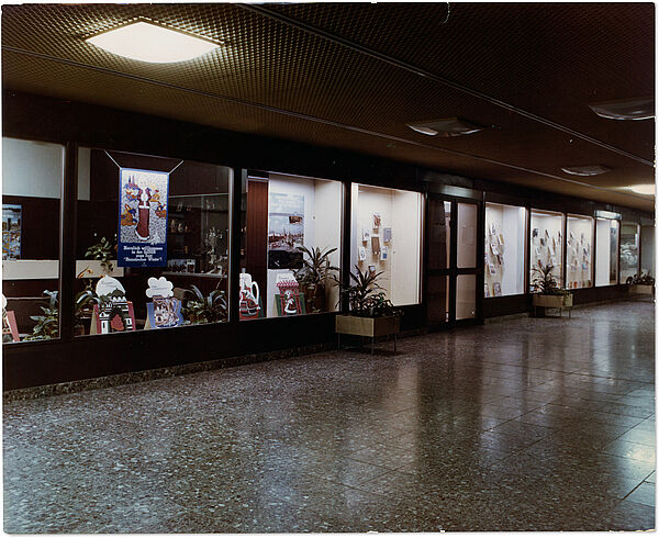 Ladenzeile des "Dienstleistungs- und Versorgungstrakts" im "Haus 18" der Stasi-Zentrale, 80er Jahre; Quelle: BArch, MfS, VRD, Fo, Nr. 146, Bild 3 Blick über einen breiten, glatt gefliesten Gang im Haus 18. Auf der gegenüberliegenden Seite sind die Schaufenster von Geschäften zu erkennen.