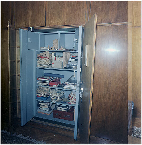 Auffindesituation der SdM-Unterlagen: Panzerschrank in einem SdM-Büro, aufgenommen während einer Durchsuchung durch die Militärstaatsanwaltschaft im Zuge der Abwicklung der Stasi, Dezember 1989; Quelle: BArch, MfS, HA IX, Fo, Nr. 610, Bild 18 Blick auf einen mannshohen, in eine Schrankwand eingebauten Panzerschrank, in dem unordentlich anmutende Unterlagen liegen.