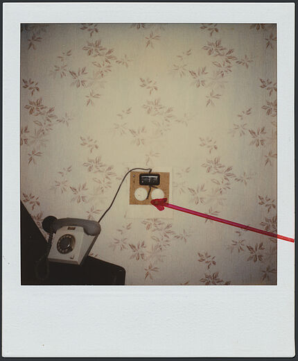 Das Polaroidfoto zeigt eine tapezierte Wand, vor der ein Telefon steht. Neben dem Telefon ist eine elektronische Apparatur zu sehen. Diese wurde auf dem Polaroidfoto mit einem roten Pfeil markiert.