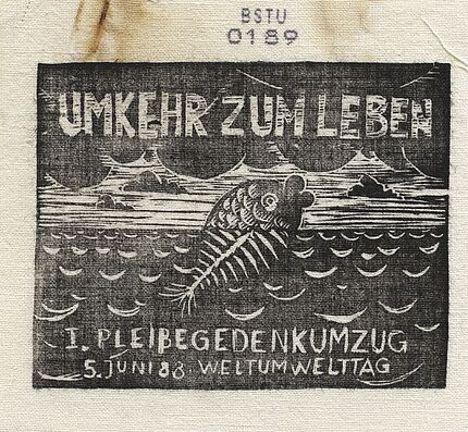 Bedrucktes Stoffstück, auf dem ein Fisch abgebildet ist, der aus dem Wasser herausschaut. Der untere, sich im Wasser befindliche Teil des Fisches besthet nur aus Gräten. Darüber steht der Text "Umkehr zum Leben", darunter "1. Pleißegedenkumzug, 5. Juni '88, Weltumwelttag